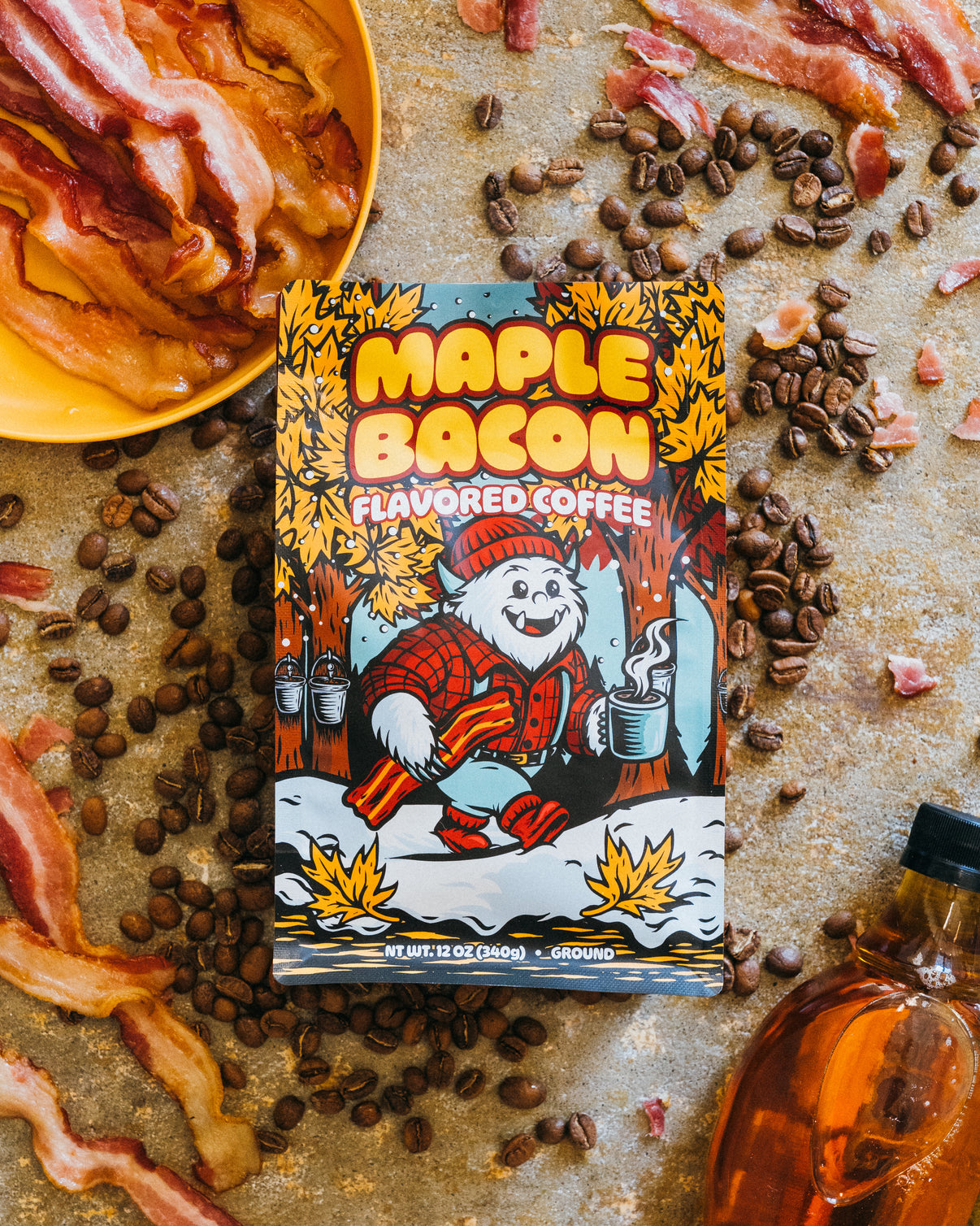 Maple Bacon Flavored Coffee package surrounded by coffee beans and bacon on a rustic surface